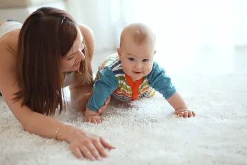 A smiling mother and her baby playing on a white fluffy carpet.